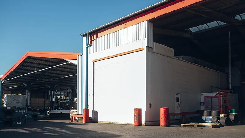 Two industrial warehouse buildings with red cladding and metal roofs stand side by side under a clear blue sky. The area between them is paved, and various equipment and pallets can be seen near the buildings.