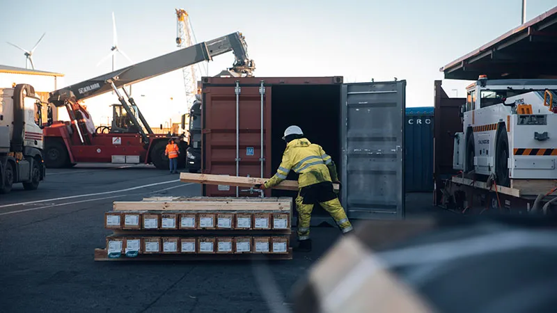 A worker in a yellow safety suit and helmet moves a wooden beam near stacked pallets in a busy shipping port, with freight containers, cranes, and vehicles in the background.