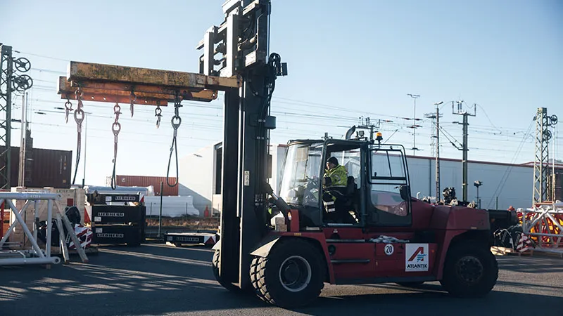 A large red forklift lifts a heavy metal beam with chains in an industrial area, while a worker operates the vehicle. In the background, cargo containers and power poles are visible. 