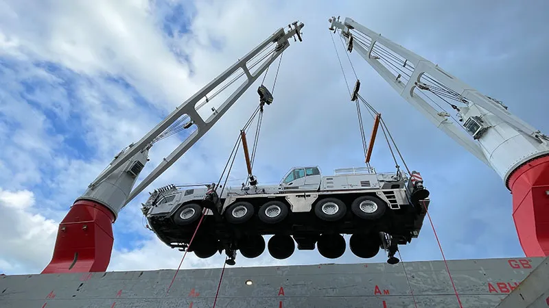 A large mobile crane truck is being lifted high above a ship’s deck by two heavy-lift ship cranes, while the sky is partly cloudy.