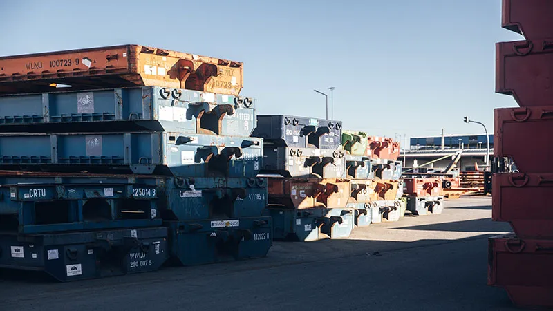 On a sunny day, large, colorful shipping containers are lined up outdoors, casting shadows on the ground with industrial facilities visible in the background.