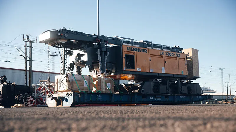 A large yellow Liebherr crane component LR 1300.1 SX is secured on a low-loader and ready for transport at an industrial site with buildings, power lines, and railway tracks in the background.