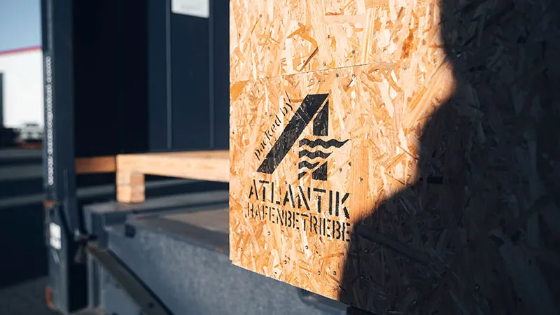 Close-up of a wooden transport crate with the ATLANTIK logo and text, standing on a loading ramp and illuminated by the sun.