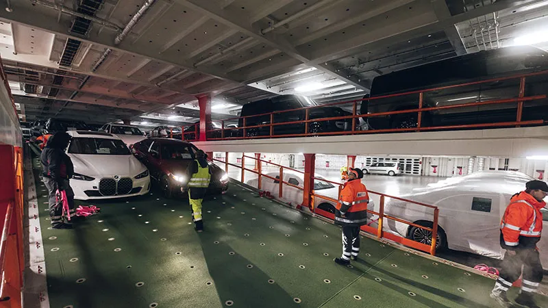 Erfahrung Arbeiter in Warnkleidung sichern Autos auf einem zweistöckigen Fahrzeugdeck im Inneren eines großen Schiffes, auf dem verschiedene Autos dicht an dicht bei künstlicher Beleuchtung geparkt sind.