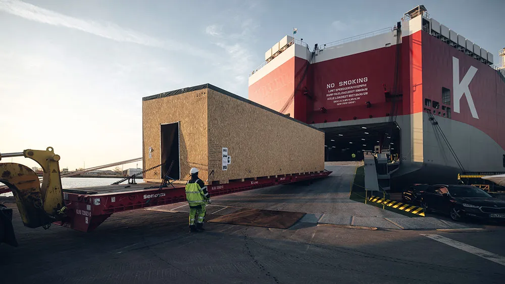 Eine große rechteckige Kiste wird in einem Hafen über einen Tieflader auf ein Frachtschiff verladen, wobei ein Arbeiter in Warnkleidung den Vorgang überwacht.