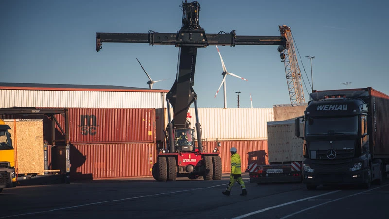Know How Ein Arbeiter in einem Warnschutzanzug überquert ein Versandgelände, wo ein großer Kran einen Container bewegt. Im Hintergrund sind bei klarem Himmel gestapelte Schiffscontainer, ein Lastwagen und Windkraftanlagen zu sehen.