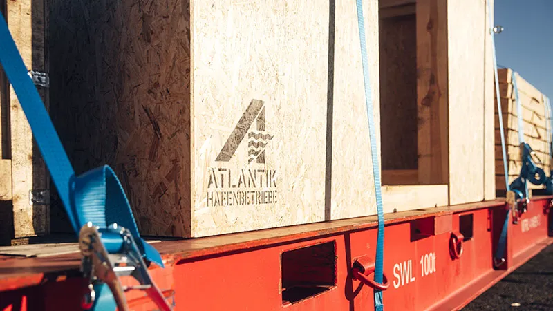 Lösungen aus einer Hand Holzkisten mit dem Logo der Atlantik Hafenbetriebe sind auf einem roten Tieflader mit blauen Gurten für den Transport bei strahlendem Sonnenschein gesichert.