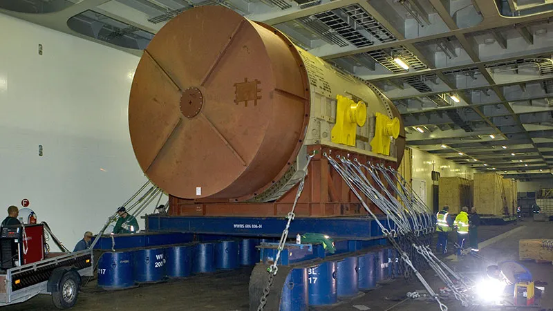A large, cylindrical industrial component is secured on a blue platform in a spacious cargo area. Several workers wearing protective gear are present, and chains anchor the component in place. The surroundings are industrial and well lit.  