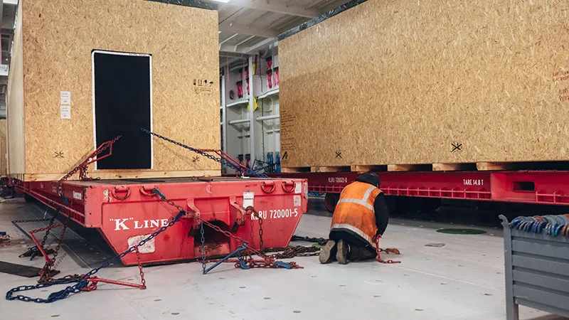 A worker in an orange safety vest kneels on the ground and secures large wooden crates with chains on red flatbed containers in a sprawling industrial facility.
