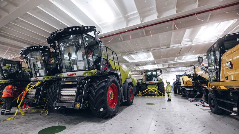 In a brightly lit, spacious industrial facility, several large agricultural and harvesting machines are parked and inspected by workers in protective clothing. Yellow tape cordons off the areas around the machines.