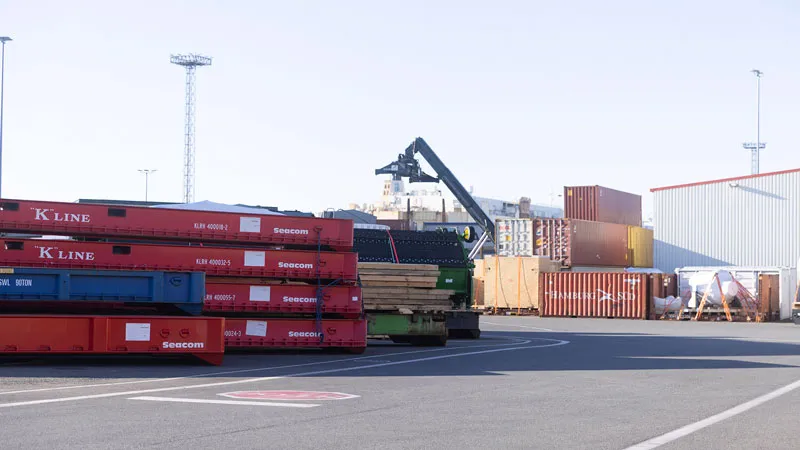A shipping yard with stacked red and blue shipping containers, a crane lifting cargo, and various shipping containers in the background near a warehouse building.