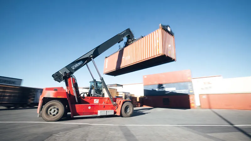 A red forklift lifts a large shipping container in a shipping yard, with other stacked containers and a clear blue sky in the background.